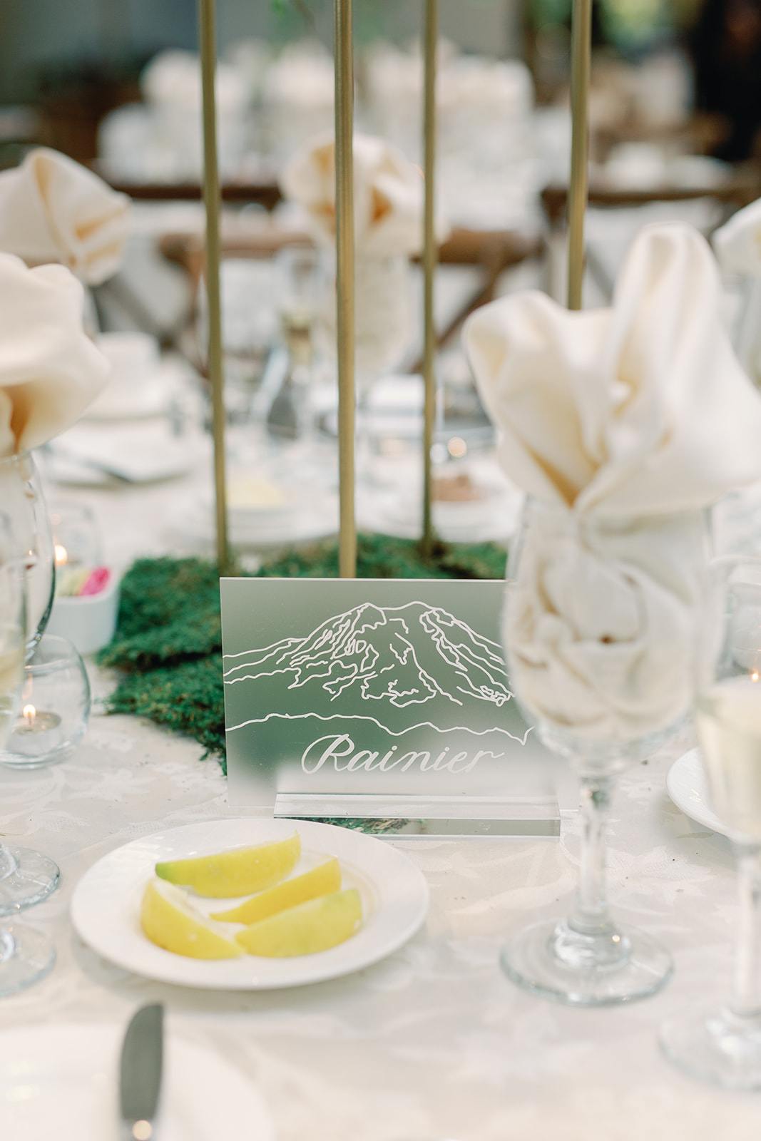 Frosted acrylic Mount Rainier table name card with mountain outline, surrounded by moss decor, sliced lemons, and napkin-filled wine glasses at Hershey Gardens wedding reception in Hershey, Pennsylvania.