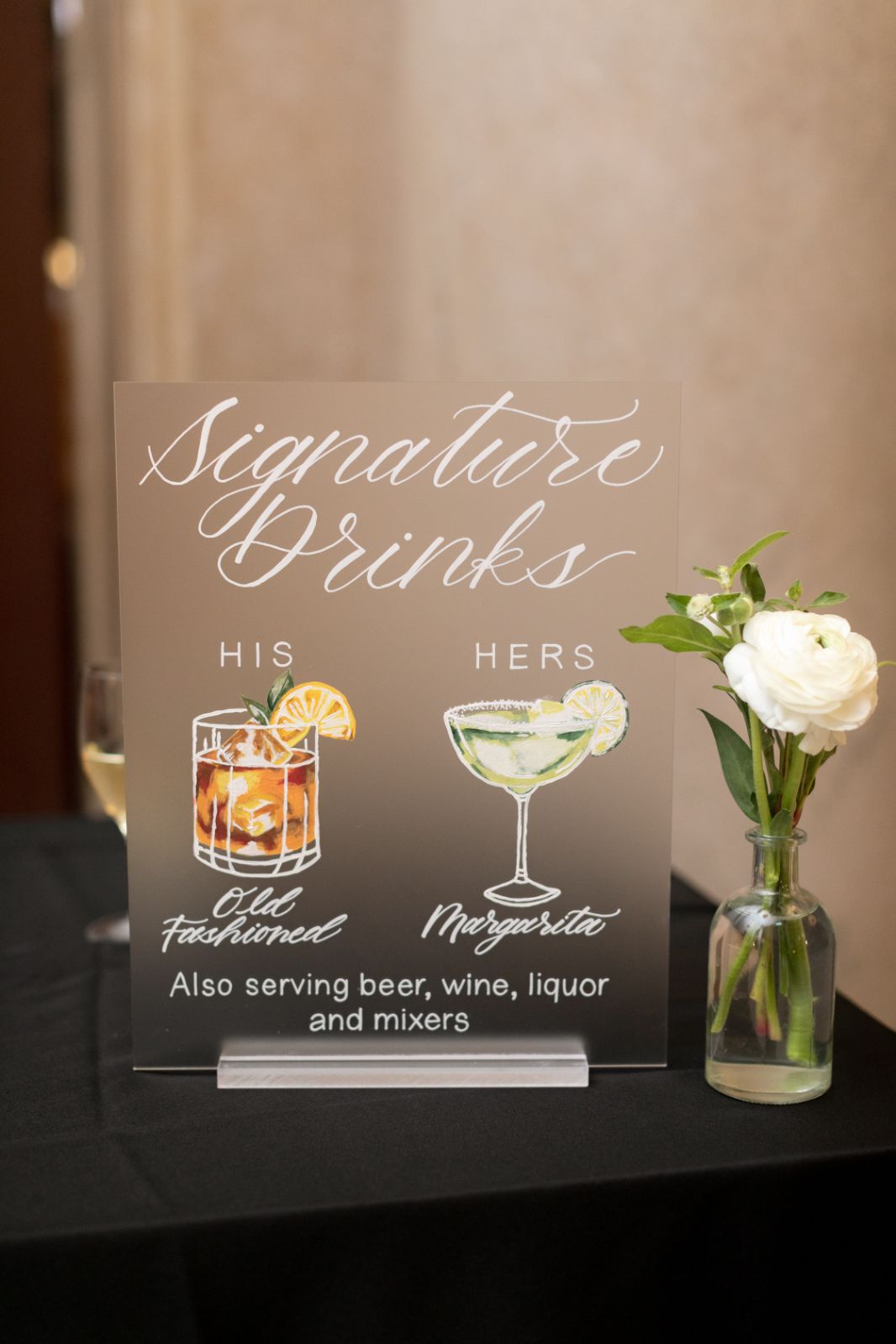 Frosted acrylic signature drinks sign with illustrated his and hers cocktails—Old Fashioned and Margarita—displayed with white rose floral vase at Free Library of Philadelphia wedding reception.