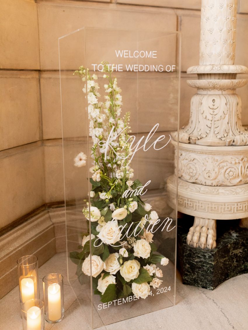 Modern clear acrylic wedding welcome sign with white and green floral arrangement and candles at the base, displayed at the Free Library of Philadelphia wedding venue.