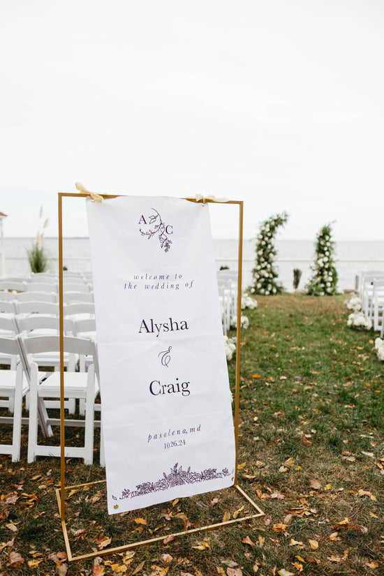 White fabric wedding welcome sign with black script, hanging on a gold stand. Surrounded by white ceremony chairs and white-and-green floral pillars at the altar. Outdoor waterfront wedding at Celebrations at the Bay in Pasadena, Maryland