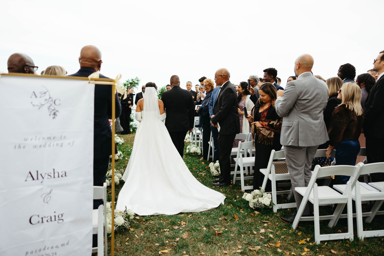 Elegant outdoor wedding ceremony at Celebrations at the Bay in Pasadena, Maryland, featuring custom fabric welcome sign and bride walking down the aisle with her father, surrounded by guests