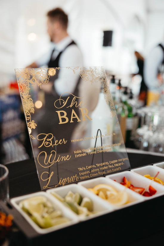 Clear acrylic bar sign with elegant gold lettering listing beer, wine, and liquor options. Bartender blurred in the background. Fresh fruit garnishes displayed in front for cocktails. Wedding reception at Celebrations at the Bay in Pasadena, Maryland.