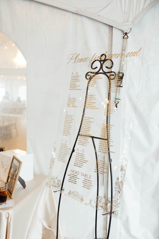 Elegant clear acrylic wedding seating chart with gold calligraphy and floral design, displayed on black easel at Celebrations at the Bay wedding reception in Pasadena, Maryland