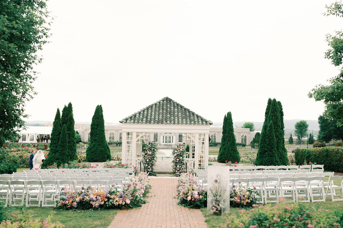 Bridgerton-style wedding at Hershey Gardens with lush pastel aisle florals, white ceremony chairs, gazebo backdrop, and acrylic floral welcome sign in Hershey. Pennsylvania.