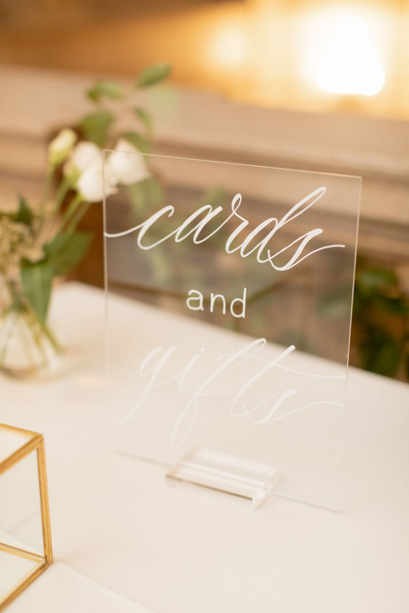 Clear acrylic cards and gifts sign with white script lettering on display at Free Library of Philadelphia wedding venue, styled with soft floral accents.
