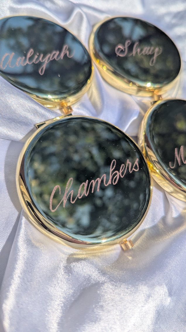 Gold compact mirrors hand engraved with the name “Chambers” in elegant script, photographed on satin fabric. Perfect for personalized bridesmaid gifts, wedding favors, or luxury event keepsakes.