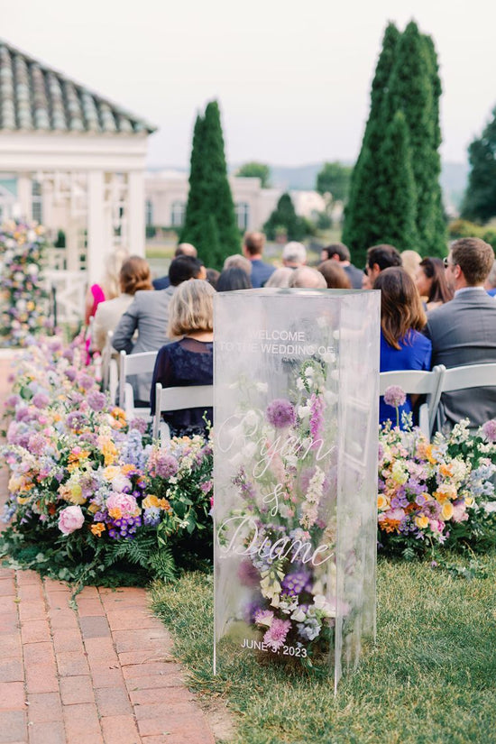 Clear acrylic box wedding welcome sign with elegant white calligraphy and pastel florals inside, surrounded by lush aisle and altar arrangements at a Bridgerton-themed ceremony at Hershey Gardens in Hershey, PA.
