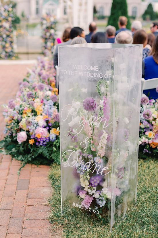Clear acrylic box wedding welcome sign with elegant white calligraphy and pastel florals inside, surrounded by lush aisle and altar arrangements at a Bridgerton-themed ceremony at Hershey Gardens in Hershey, PA.