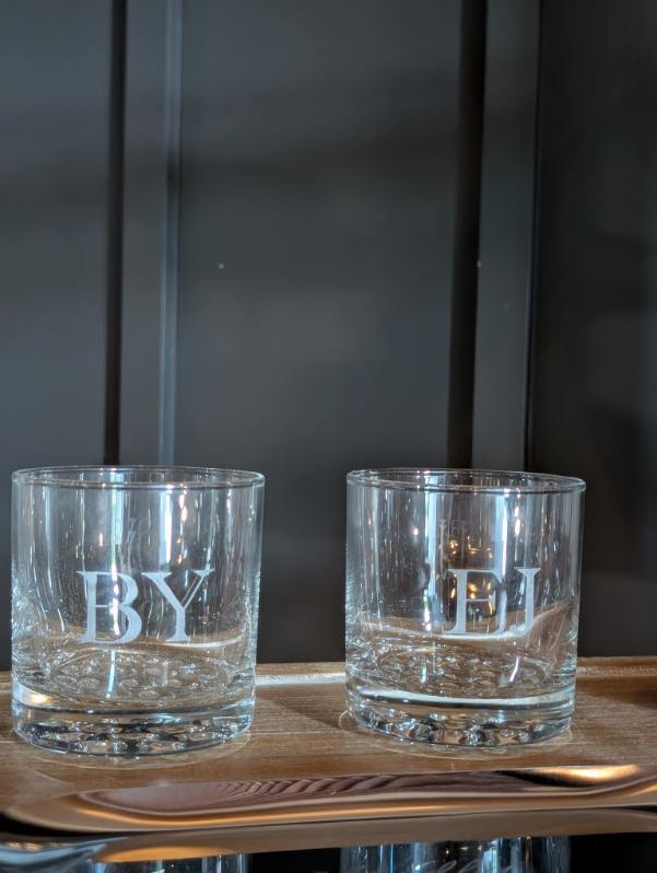 Two hand engraved whiskey glasses personalized with initials "BY" and "EJ" in a clean sans serif font at a wedding reception at The Ivy at Ellis Preserve in Philadelphia, PA. Custom live wedding engraving by SLM & Design Co.