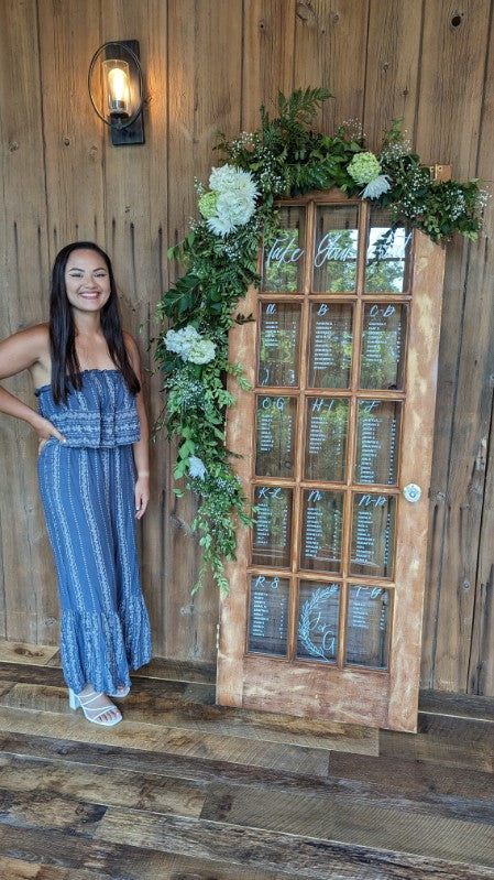 Calligrapher standing beside hand-lettered wooden glass panel seating chart adorned with green and white florals at Domaine Pterion Winery and Vineyard wedding in Macungie, PA.