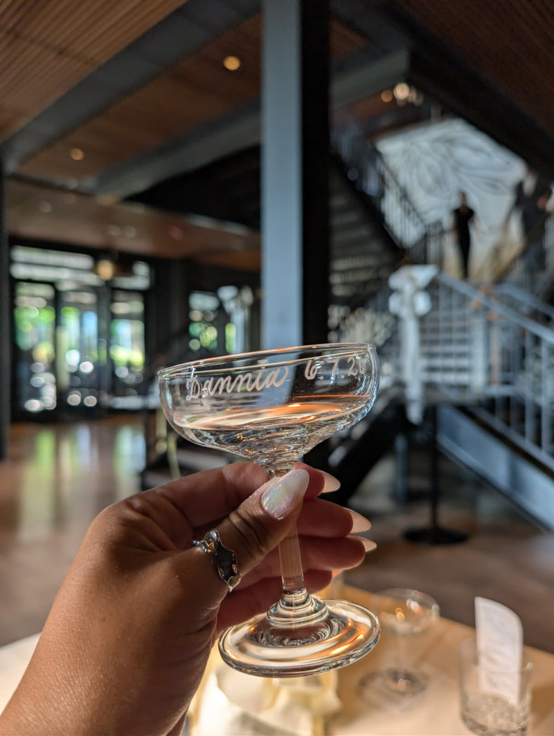 Live engraved coupe glass personalized with the name "Dannia" and wedding date "6.7.26" during a wedding reception at The Ivy at Ellis Preserve in Philadelphia, PA. Custom wedding guest keepsake engraved on-site by SLM & Design Co.