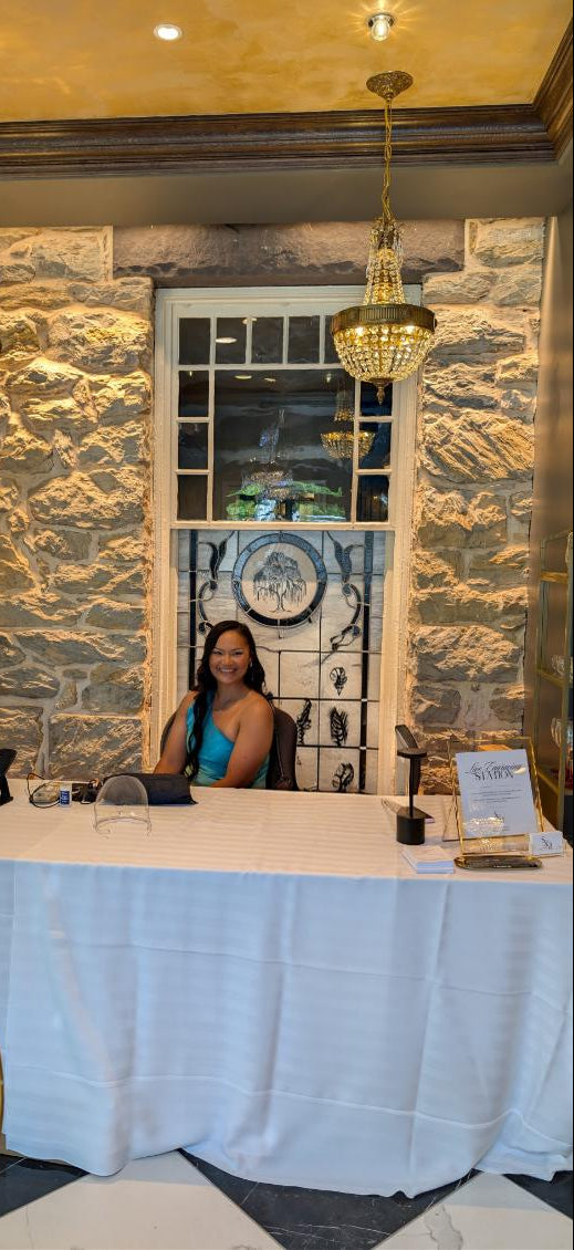 Tiffany of SLM & Design Co. seated at a cocktail hour setup surrounded by glassware favors and engraving tools, performing live engraving at a wedding reception at The Willows at Ashcombe in Mechanicsburg, PA. Custom on-site wedding personalization experience.