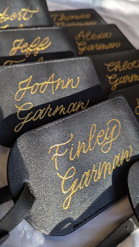 Black leather luggage tags hand heat foiled with gold metallic lettering, featuring personalized names in elegant script with name "Finley Garman." Perfect for luxury travel gifts, weddings, brand activations, corporate events and custom event favors.