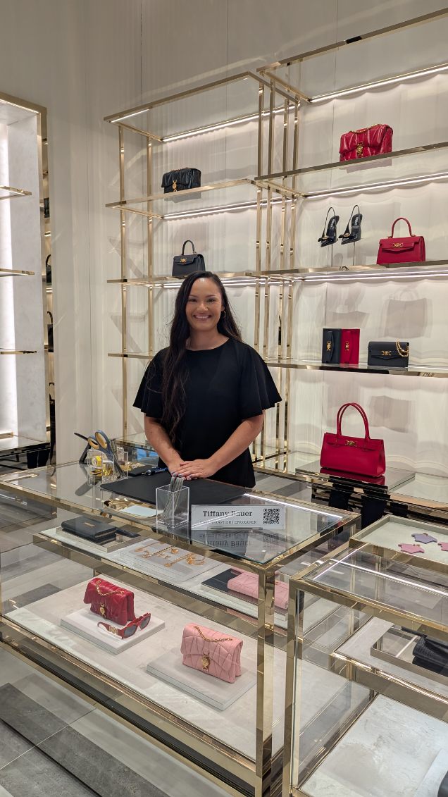 Luxury brand activation for Mother’s Day at Versace in King of Prussia, PA featuring live heat foiling by artist Tiffany Bauer. Guests received complimentary personalized red, pink, or purple leather tags with purchase. Tiffany stands in-store surrounded by luxury Versace handbags and accessories, with heat foiling supplies displayed on the counter.
